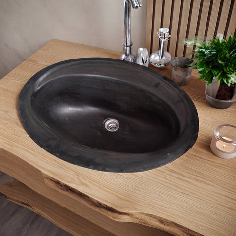 Image of An oval black stone or concrete bathroom sink basin installed into a light wood vanity countertop, with a chrome faucet and small decor items nearby.