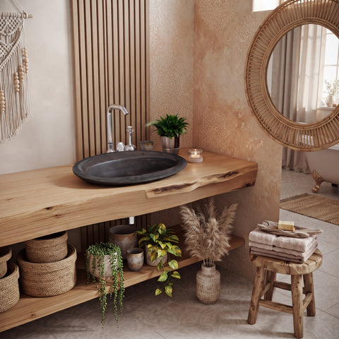 Image of A rustic wooden floating vanity with an integrated dark stone vessel sink, styled in a warm bohemian bathroom setting with plants, woven baskets, a round wicker mirror, and a small stool with folded towels.