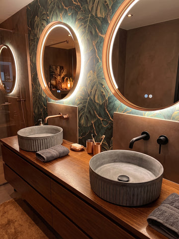 Image of A modern bathroom vanity with two round gray vessel sinks on a wooden countertop, illuminated oval mirrors with warm lighting, and a decorative green tropical leaf wallpapered wall.