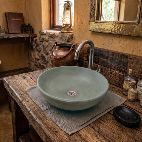 Image of A round, light sage green ceramic vessel sink basin resting on a rustic wooden vanity table with a textured gray mat. A brushed silver metal faucet is mounted on the wall behind it.