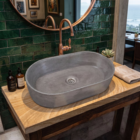 Image of A gray concrete vessel sink with a copper faucet, mounted on a wooden vanity against a green tiled wall.