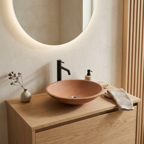 Image of A terracotta-colored ceramic vessel sink basin with a matte black faucet, set on a light oak wood vanity in a modern bathroom with a built-in circular LED mirror.