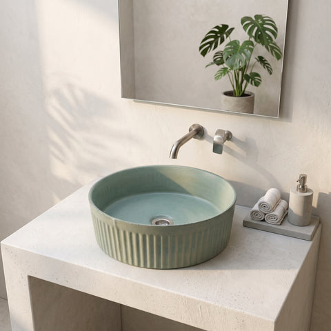 Image of A round ceramic vessel sink in a muted sage green color with vertical fluted detailing, installed on a light beige stone countertop. The scene is styled with a matching faucet, a wall-mounted mirror reflecting a potted monstera plant, and a small soap dispenser on a tray.