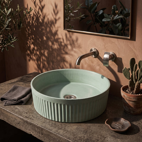 Image of A round ceramic vessel sink in a pale mint green color with vertical fluting, mounted on a concrete countertop with a matching wall-mounted faucet. The scene is styled with a folded gray towel, a small cactus in a pot, and a mirror on a textured terracotta wall.