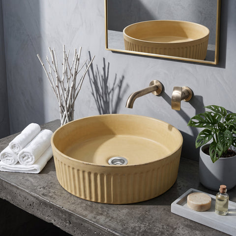 Image of A round beige ceramic vessel sink with a ribbed fluted edge, mounted on a gray concrete-look countertop with a matching wall-mounted brass faucet. A gold-framed mirror reflects the sink, and decorative props including white towels, a potted plant, and a small soap dish are arranged around it.