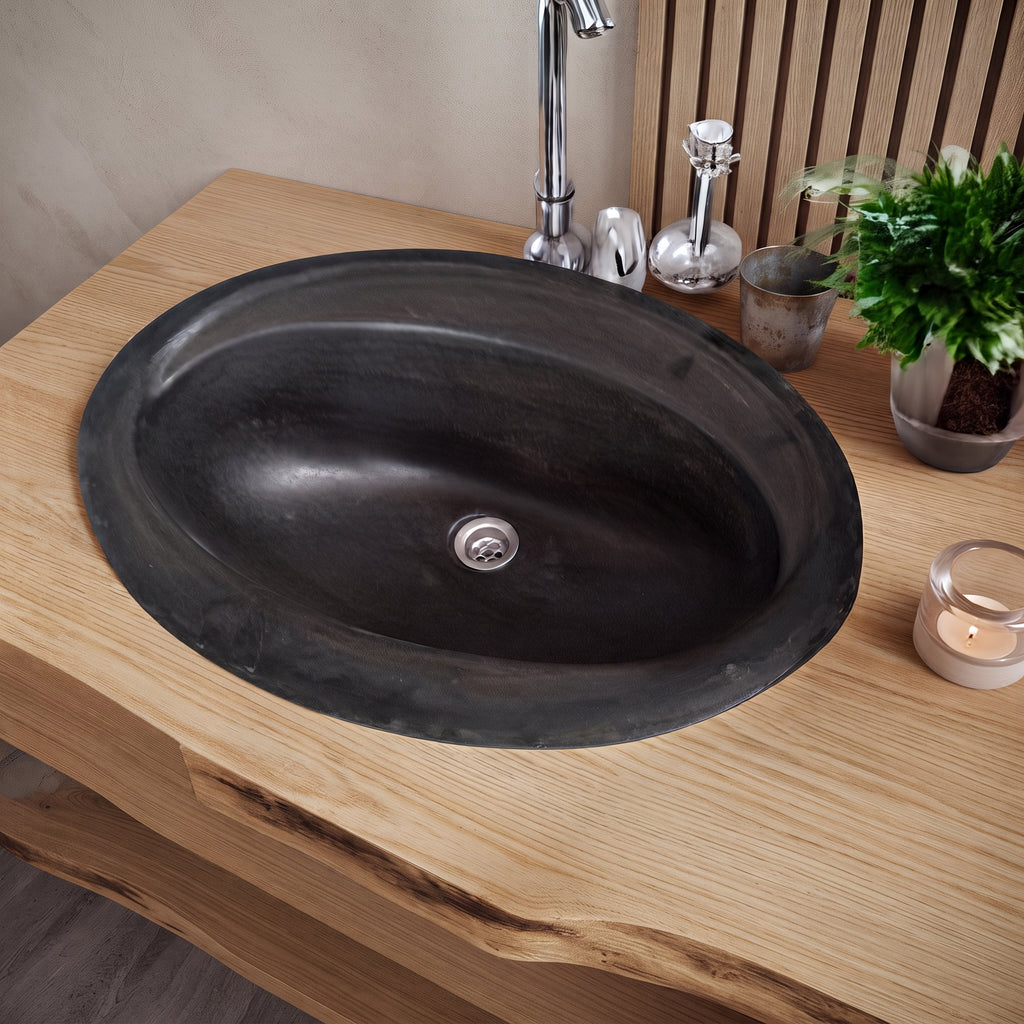 An oval black stone or concrete bathroom sink basin installed into a light wood vanity countertop, with a chrome faucet and small decor items nearby.