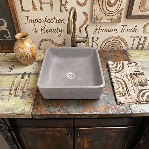 Image of A square concrete vessel sink with a matte gray finish, styled on a rustic distressed wooden countertop with a brass faucet, decorative tools, and a patterned cloth.