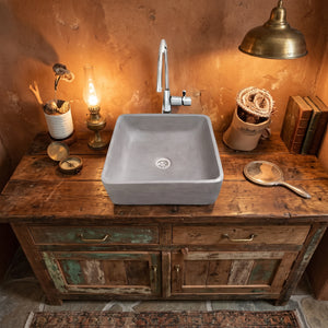 A square concrete vessel sink with a chrome faucet, mounted on a rustic distressed wooden vanity cabinet in a warm, vintage-style room with a terracotta wall, antique lamp, and books.