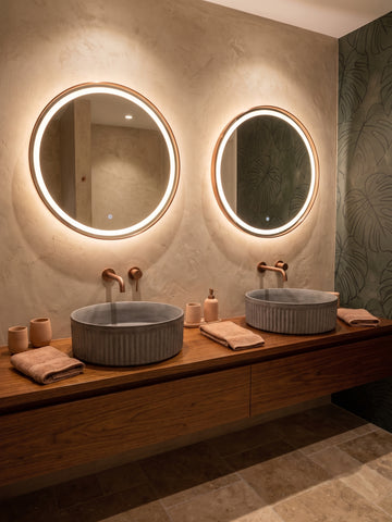 Image of A modern bathroom vanity with two round backlit mirrors, dual gray vessel sinks, warm brass faucets, and folded towels on a wooden console against a textured beige wall.