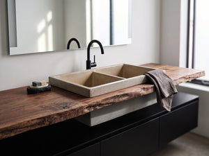 A modern double-bowl concrete farmhouse sink with a matte black gooseneck faucet, mounted on a live-edge wood vanity countertop in a bright bathroom setting.