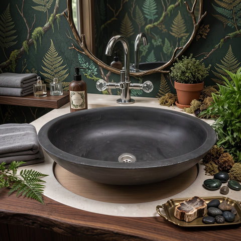 Image of A dark charcoal stone vessel sink set into a light marble countertop with a round wood inlay, styled in a bathroom with mossy fern wallpaper, a silver faucet, folded gray towels, and green plants.
