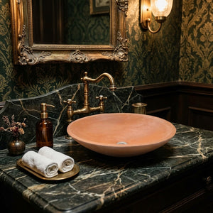 A terracotta-colored ceramic vessel sink with a matching curved brass faucet, set on a dark green marble countertop with a gold-leaf patterned wallpaper background.