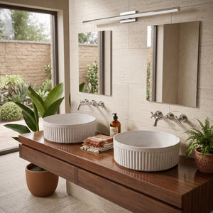 A modern double-sink bathroom vanity with two white ribbed ceramic vessel sinks on a dark wood floating console, set against beige stone tile walls with mirrors, wall-mounted faucets, and potted plants.
