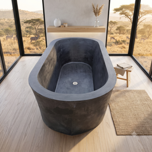 A freestanding rectangular bathtub with a dark charcoal concrete finish, positioned in a modern bathroom with large windows overlooking an African savanna landscape. A small wooden stool with books sits to the right of the tub, and a woven rug is on the floor.